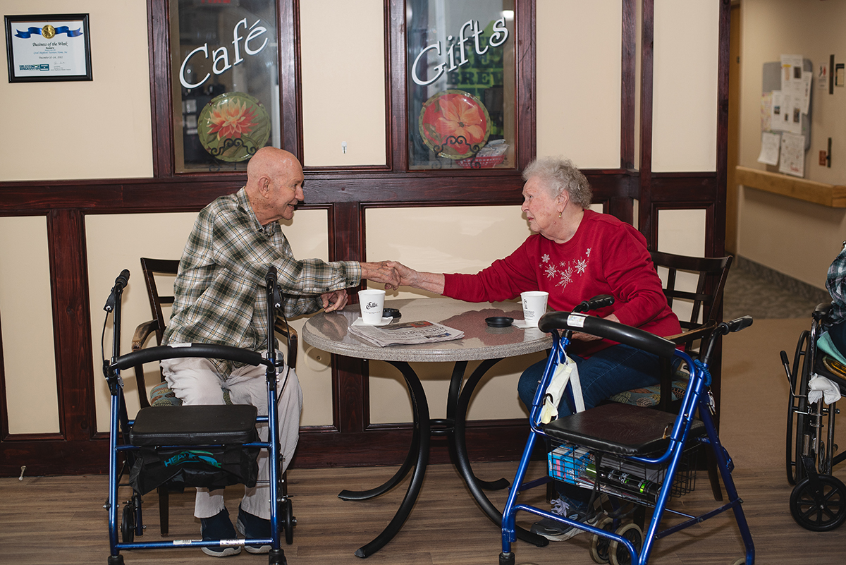 Two elderly residents smiling and holding hands at a dining table